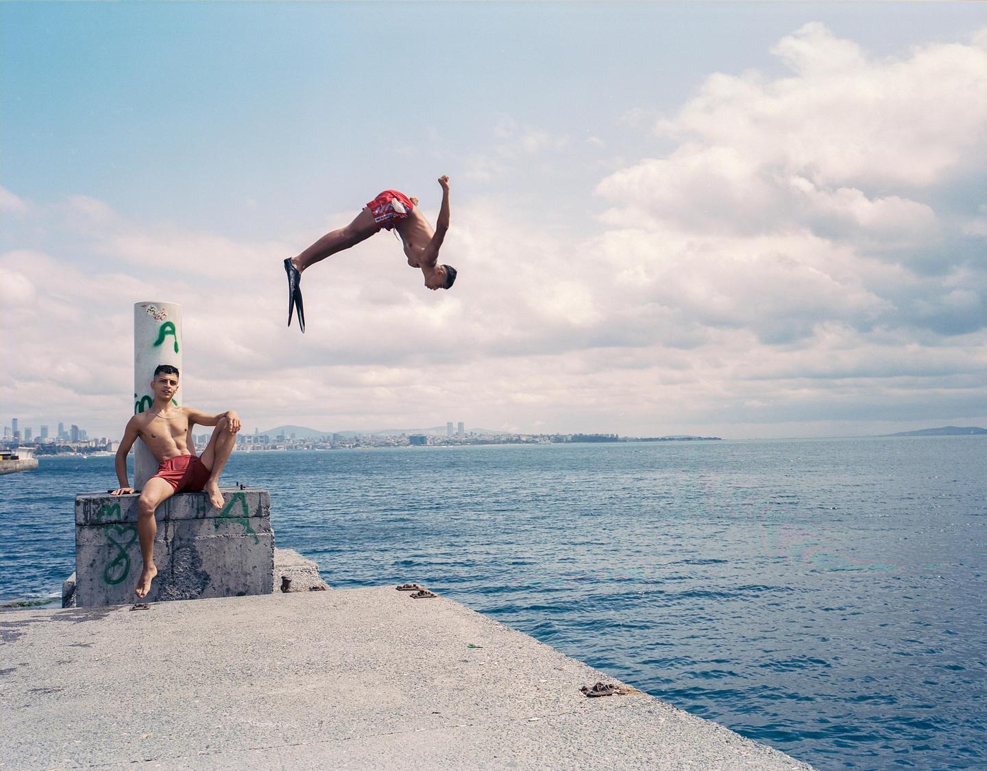 A man in red shorts sits on a concrete pier while another man in red shorts and flippers does a backflip over the water with a city skyline in the distance.