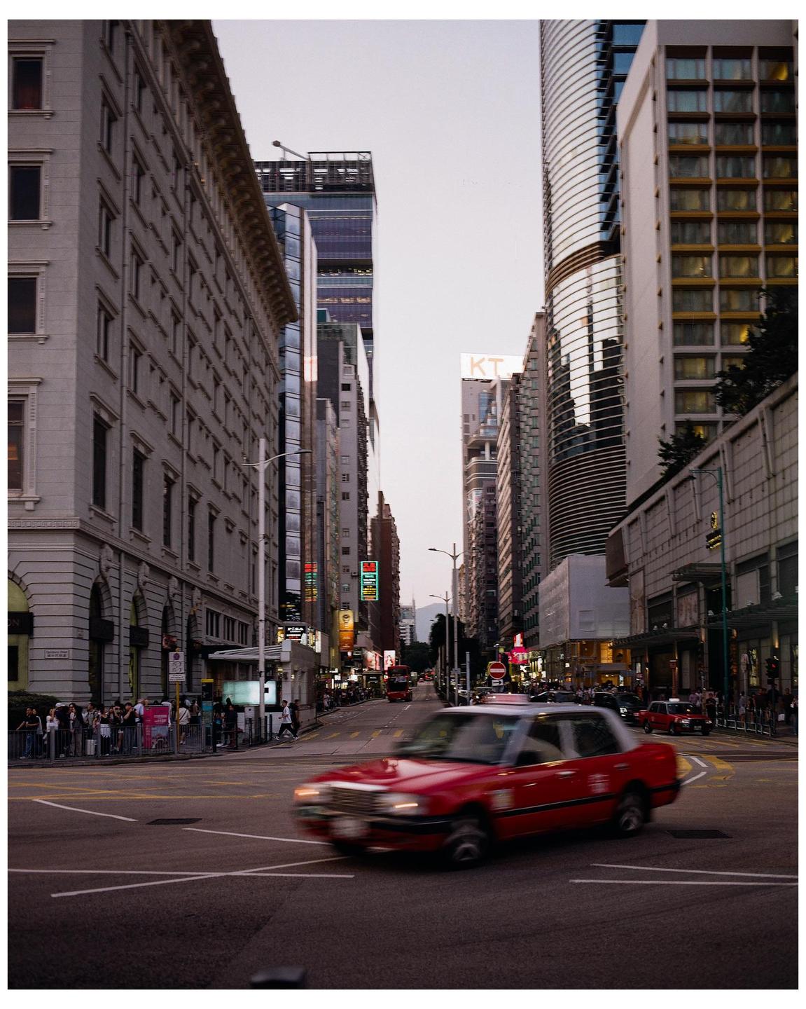 A red taxi drives through a city street lined with tall buildings and people on the sidewalk.