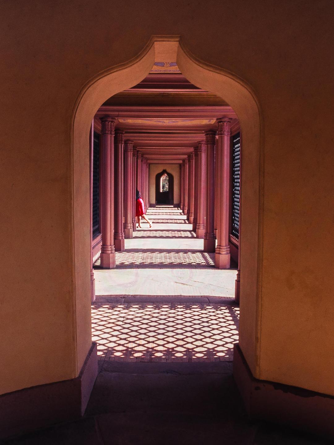 A person in a red dress walks down a long hallway with columns and patterned shadows on the floor.