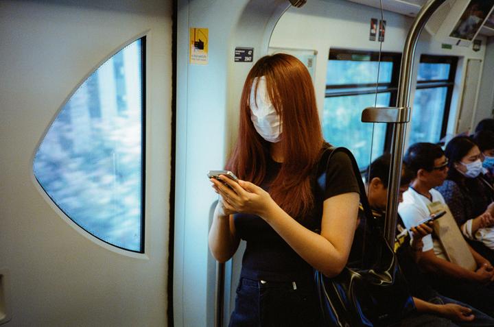 A woman with long red hair and a white face mask stands in a train car, looking at her phone.
