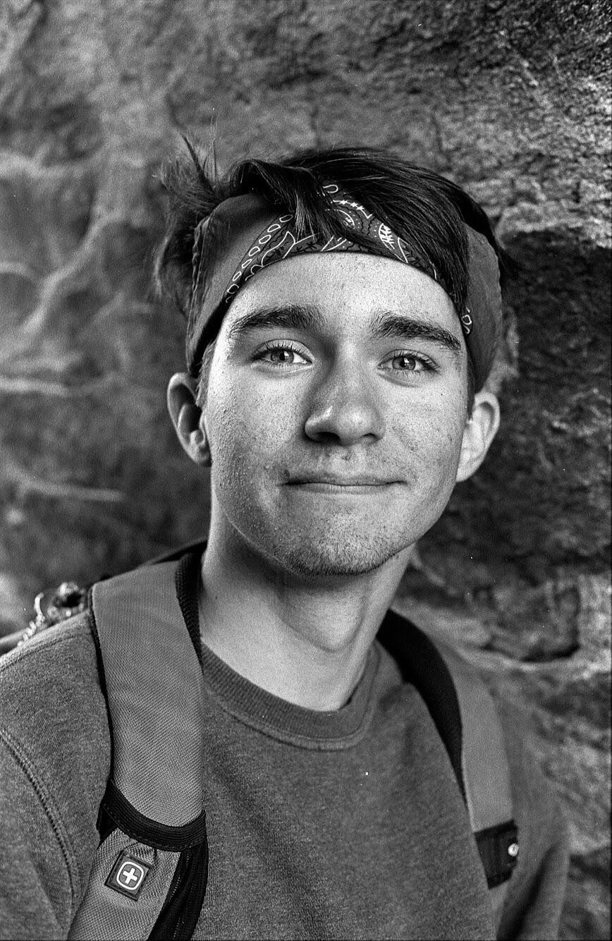 A young man wearing a bandana and backpack smiles at the camera.