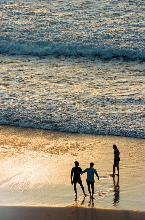 Three people stand on a beach at the edge of the ocean as waves roll in.