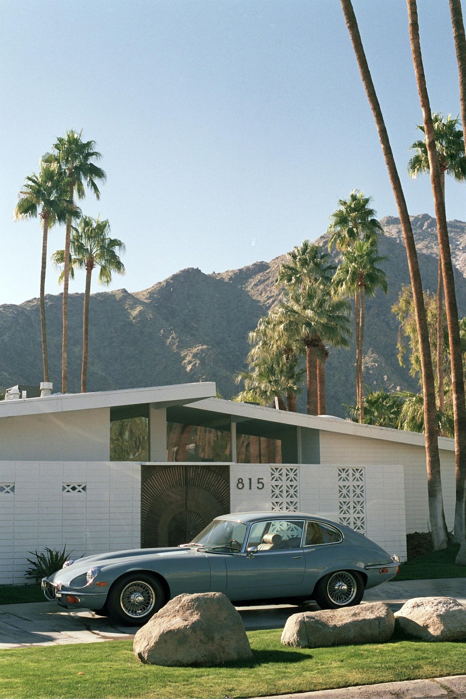 A blue car is parked in front of a white house with palm trees and mountains in the background.