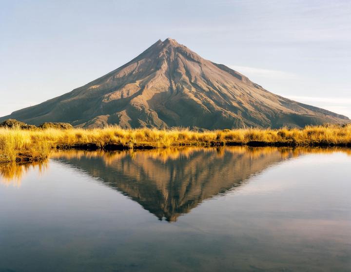 A mountain is reflected in a still body of water, with golden grass in the foreground.