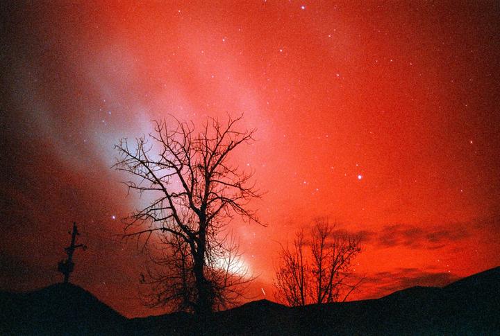 A vibrant red night sky filled with stars and a faint aurora borealis glows above silhouetted hills and bare trees.