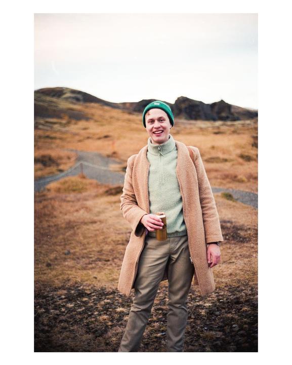 A man in a green beanie and a brown coat smiles while holding a can in a barren, hilly landscape.