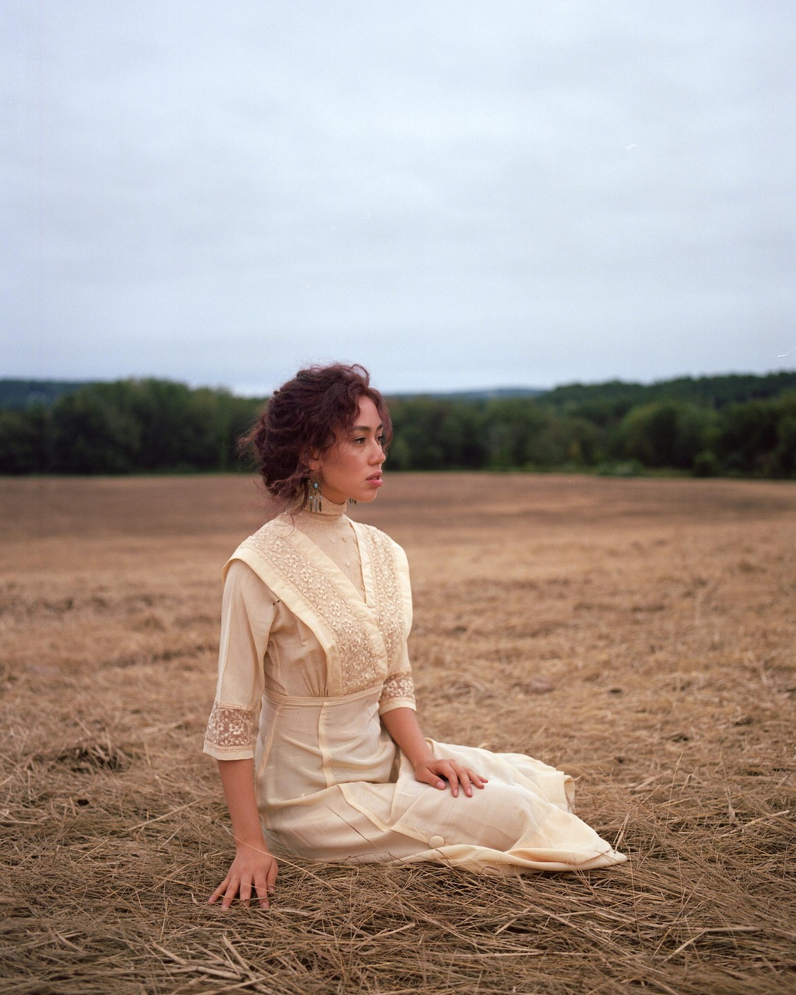 A woman in a vintage dress sits in a field of dry grass under an overcast sky.