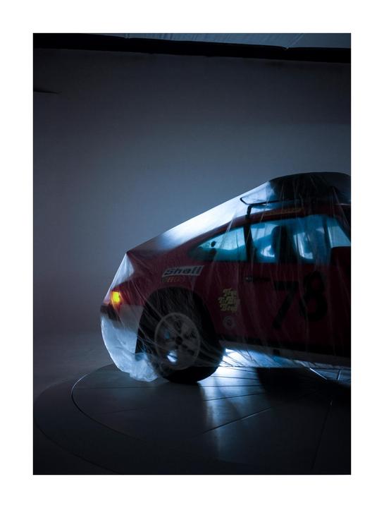 A red toy car covered in plastic is illuminated in a dark room.
