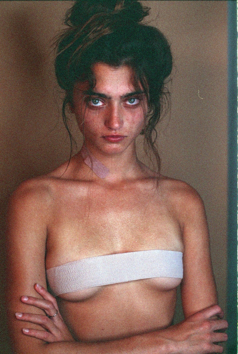 A woman with dark hair and intense eyes is shown from the chest up, with a bandage wrapped around her chest and another on her neck.