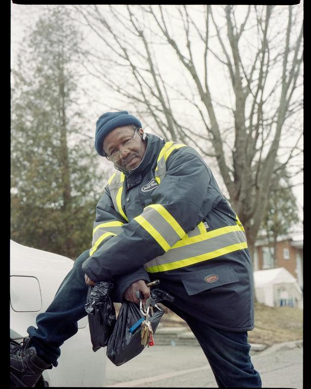 A man wearing a blue beanie and a reflective jacket is leaning on a white car while holding two black bags and a set of keys.