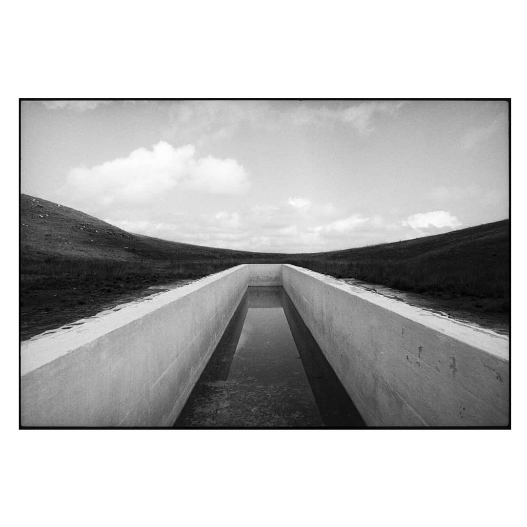 A concrete channel filled with water stretches into the distance between two grassy hills under a cloudy sky.
