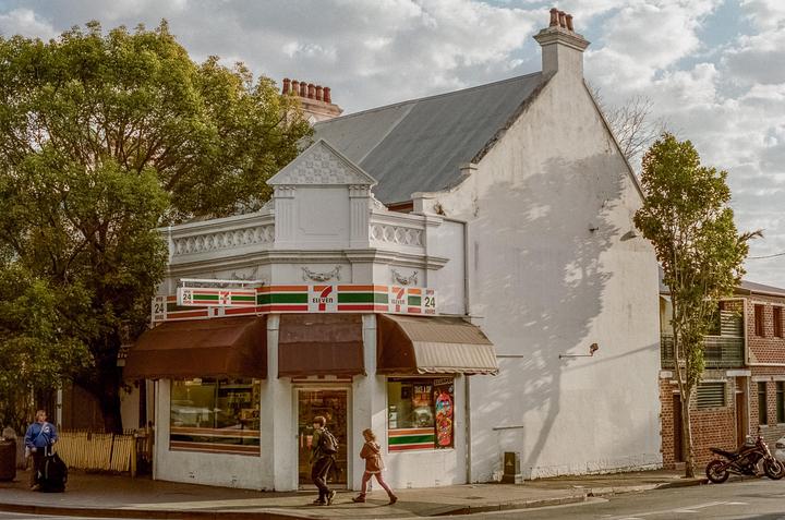 A white corner building with a 7-Eleven store on the ground floor is visible from the street, with trees and people walking by.