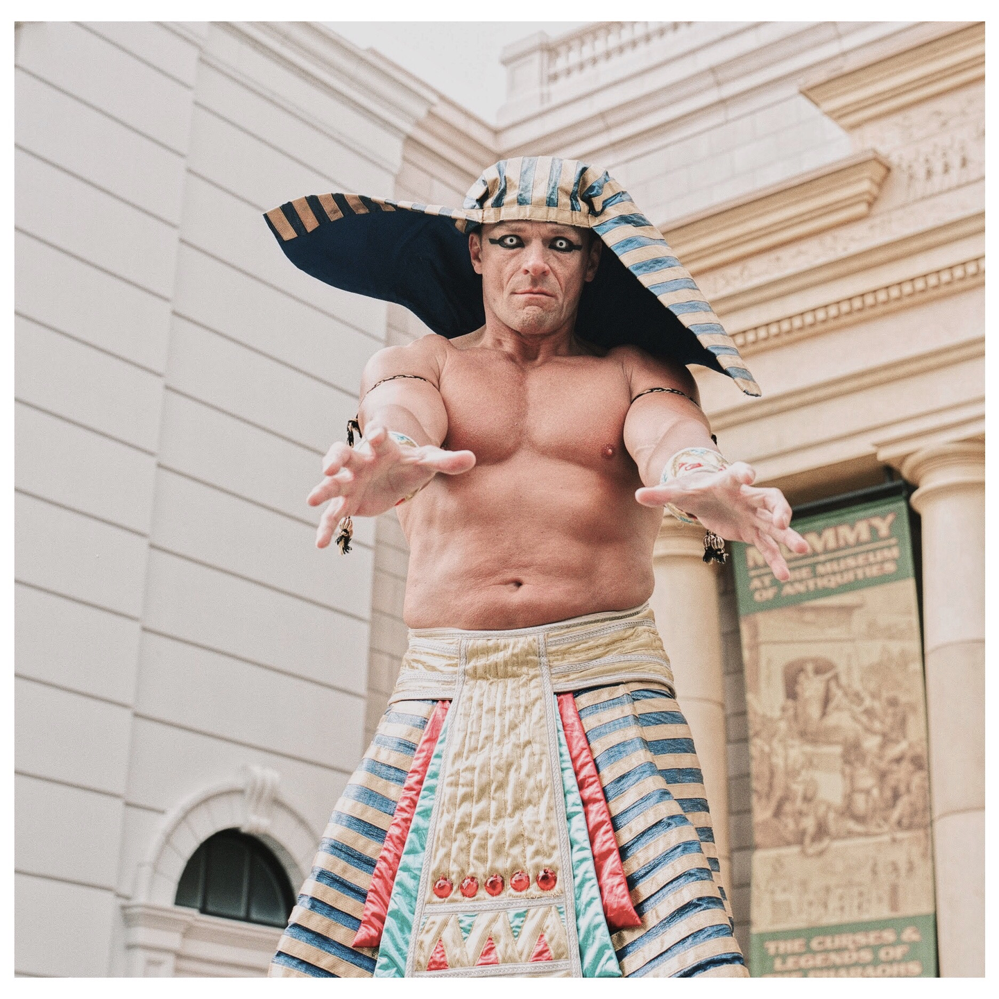 A man in an elaborate costume with white contact lenses and outstretched arms stands in front of a building.