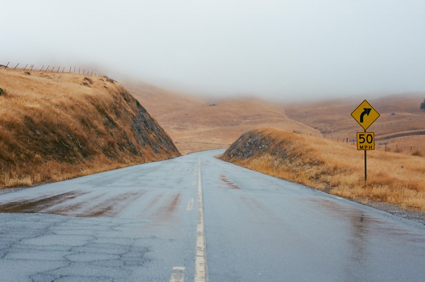 A wet road curves through golden hills under a hazy sky, with a yellow road sign indicating a turn and a 50 mph speed limit.