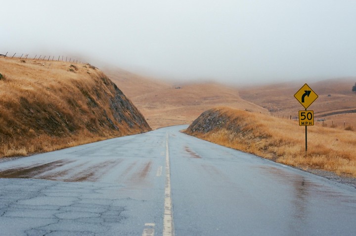 A wet road curves through golden hills under a hazy sky, with a yellow road sign indicating a turn and a 50 mph speed limit.