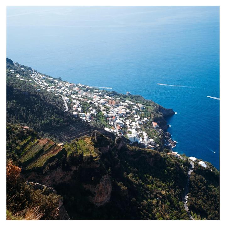 A coastal town with white buildings is nestled into a green hillside overlooking a vast blue sea.