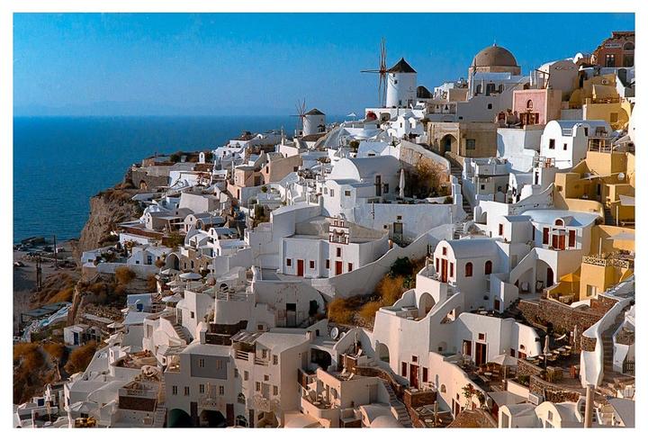 A village of white buildings with red roofs and windmills is built into a cliff overlooking the ocean.
