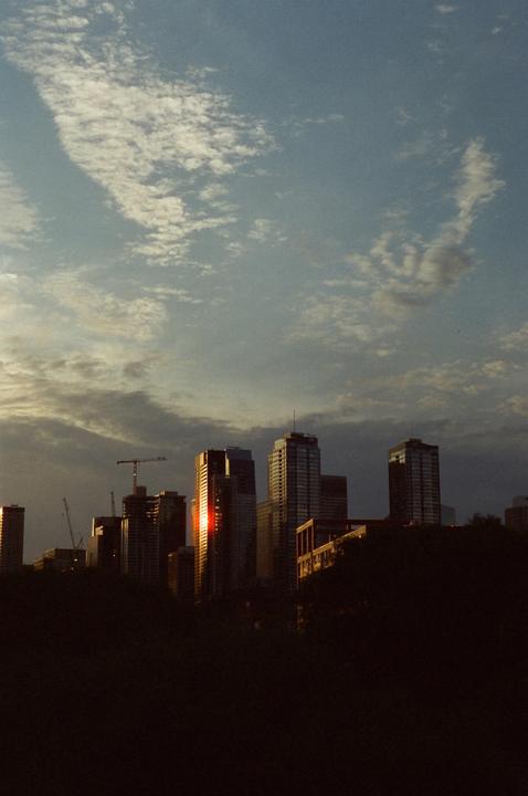 A cityscape with tall buildings is visible against a cloudy sky, with one building reflecting the setting sun.