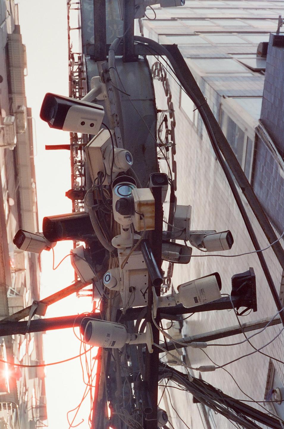A cluster of surveillance cameras and wires are mounted on a pole with buildings in the background.