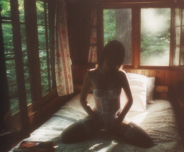 A woman kneels on a bed in a cabin, backlit by a window looking out onto a forest.