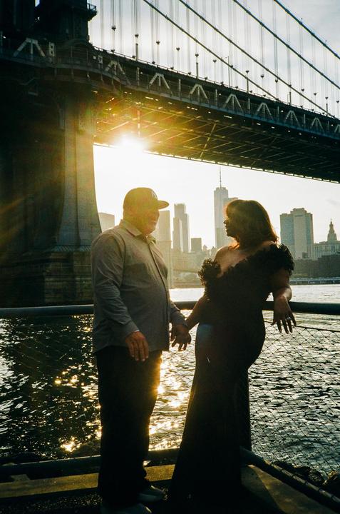 A couple stands holding hands by the water with a bridge and city skyline in the background at sunset.
