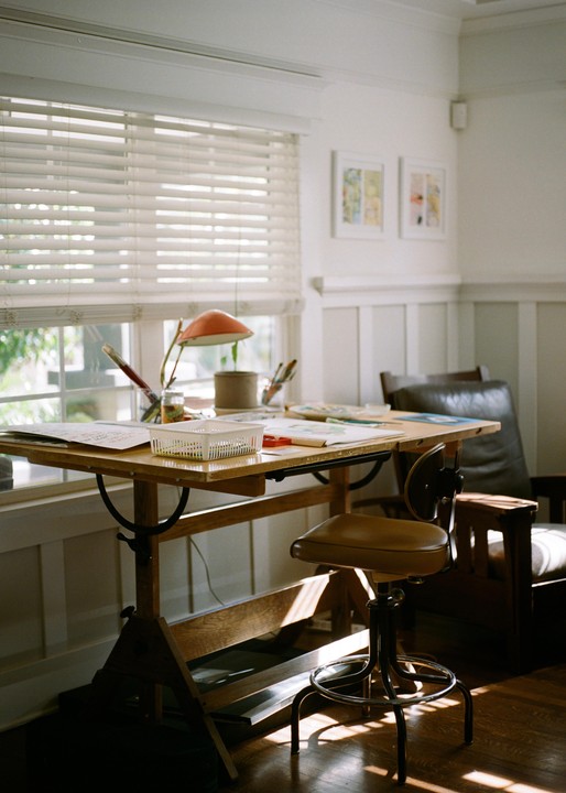 A sunlit art studio features a drafting table with art supplies, a stool, and a leather armchair next to a window with blinds.