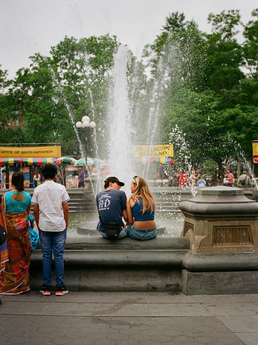 A couple sits on the edge of a fountain while two people stand with their backs to the viewer.