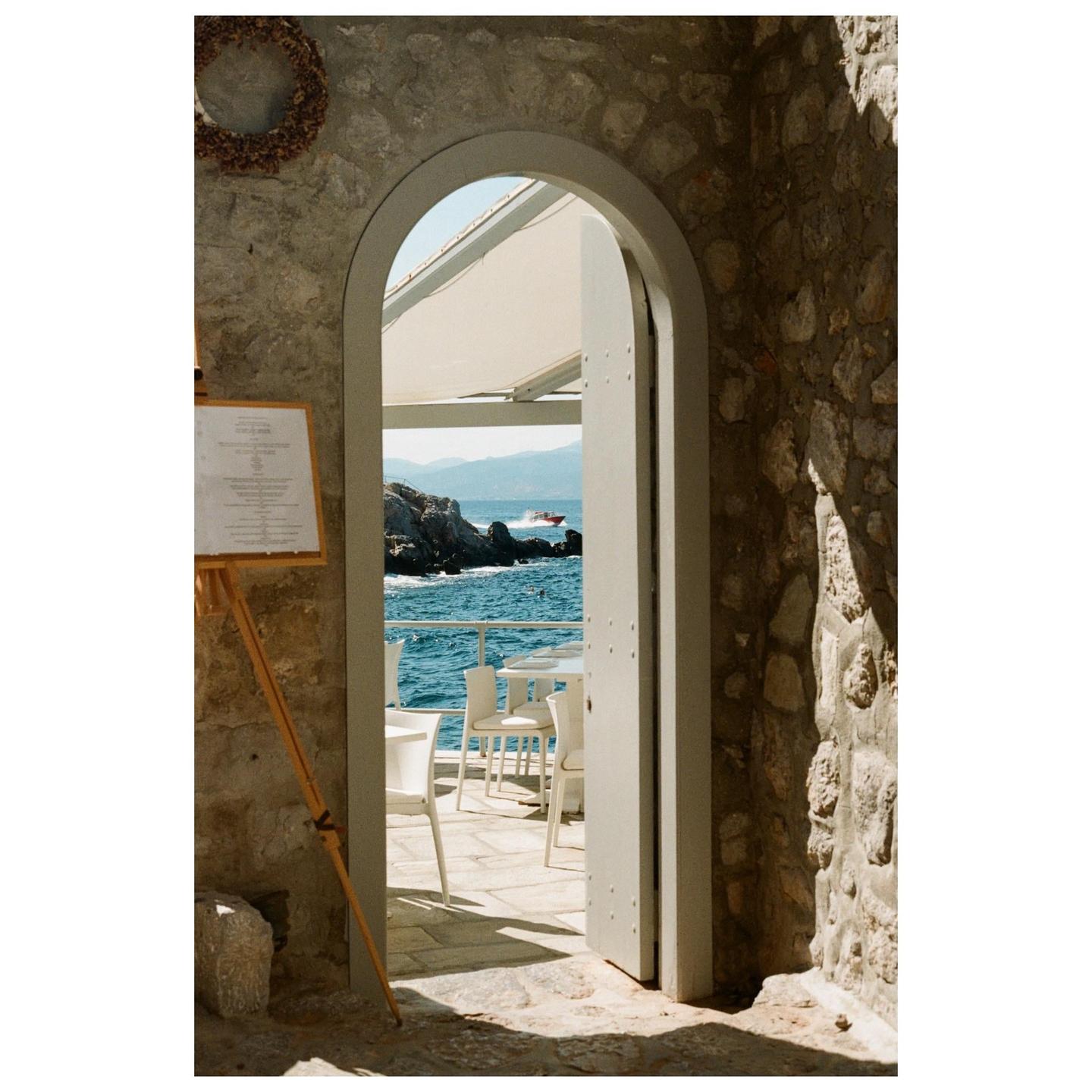 An arched doorway opens to a patio with white tables and chairs overlooking the sea with mountains in the distance.