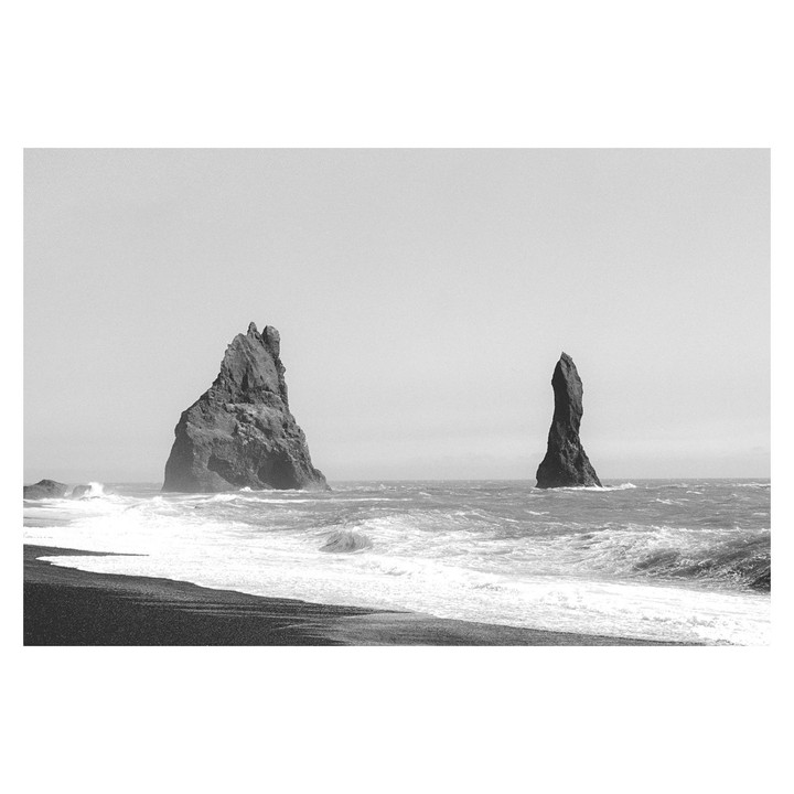 Two sea stacks rise from the ocean next to a black sand beach with waves crashing on the shore.