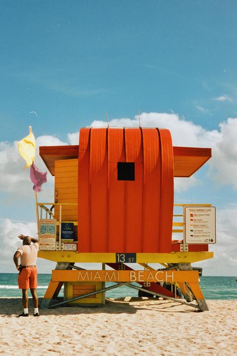 A man stands on a sandy beach looking out at the ocean next to a colorful lifeguard stand.