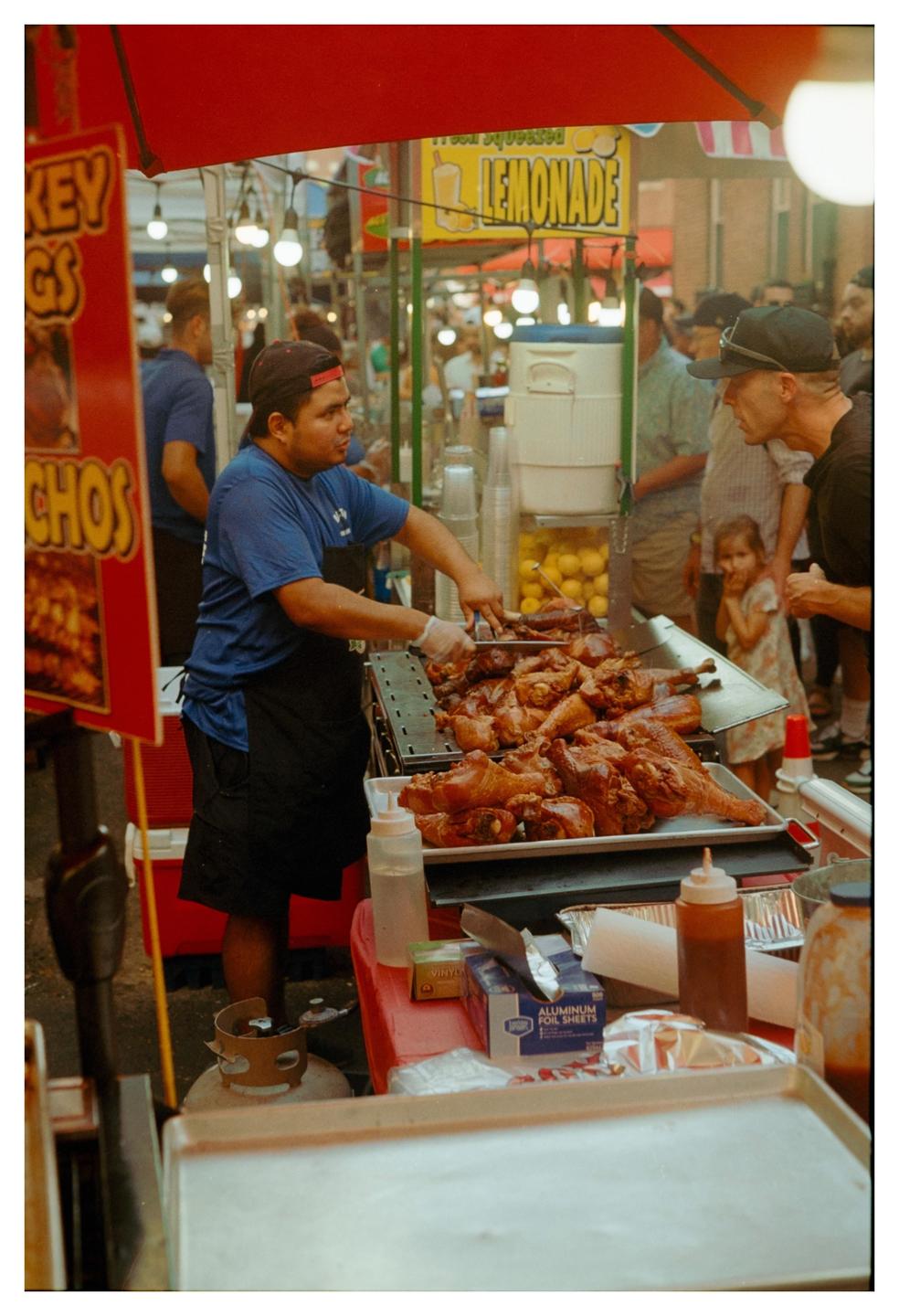 A man in a blue shirt and black apron serves roasted turkey legs from a food stall at a market.