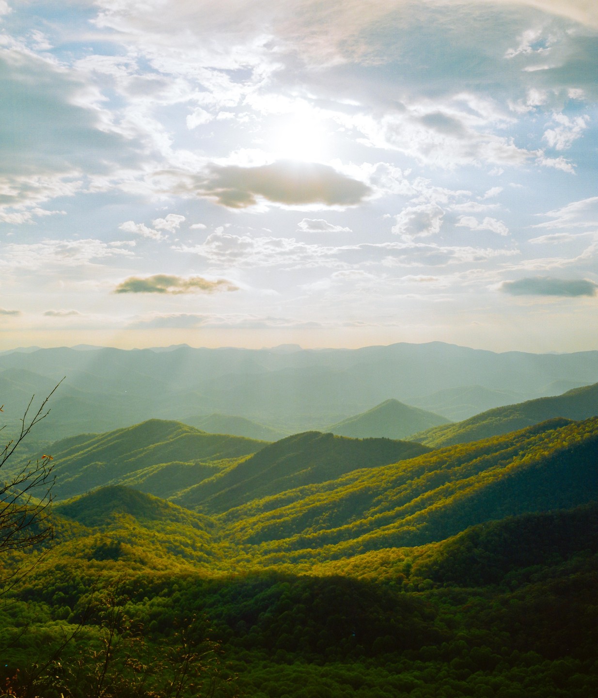 Sunlight beams through clouds onto a vast mountain landscape covered in green trees.