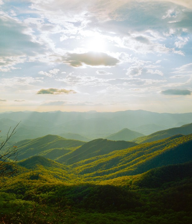 Sunlight beams through clouds onto a vast mountain landscape covered in green trees.