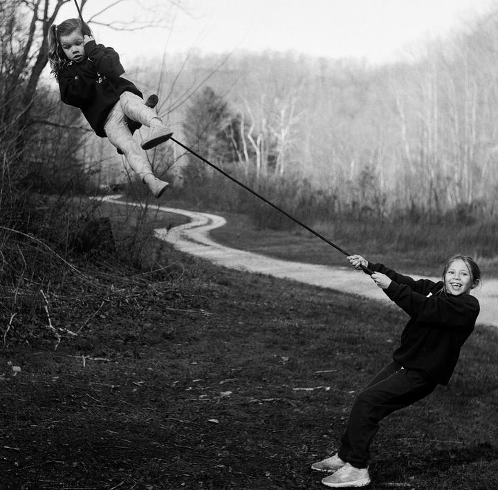 A girl pulls a rope with another girl hanging from it over a winding path in a field.