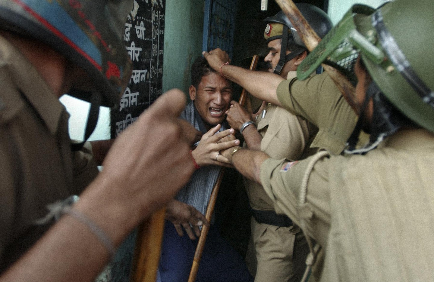 Three police officers in uniform and helmets are restraining a crying man who is holding a wooden stick.