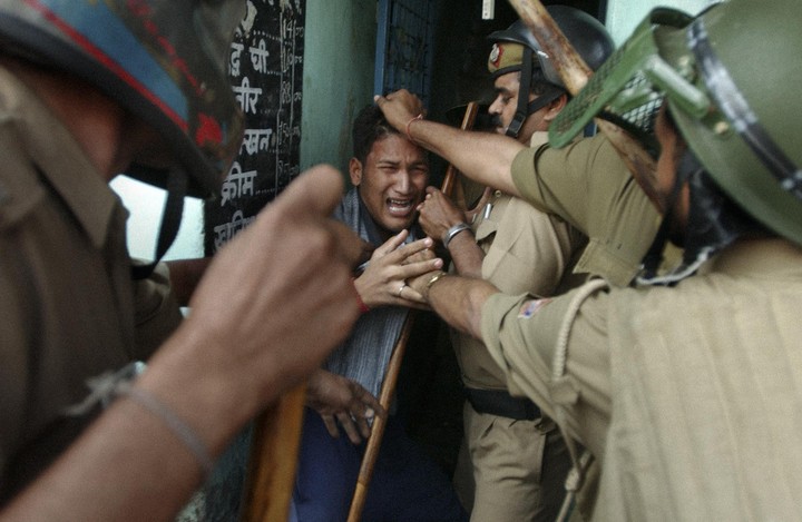 Three police officers in uniform and helmets are restraining a crying man who is holding a wooden stick.