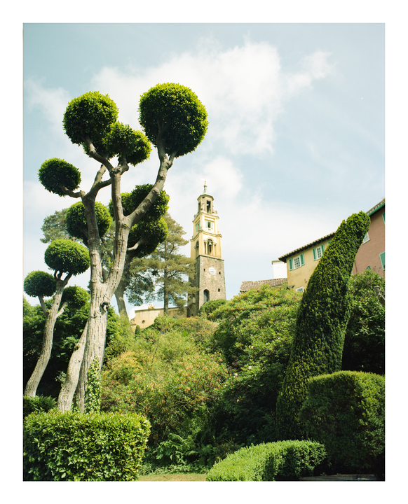 A tall clock tower rises above lush green foliage and buildings under a partly cloudy sky.