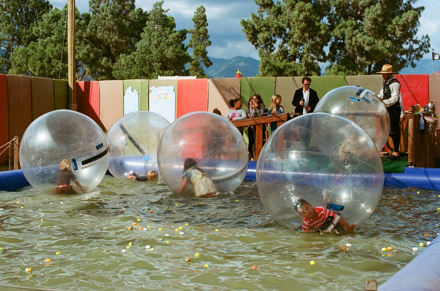 Children play in large inflatable balls in a pool of water while adults watch from a wooden platform.