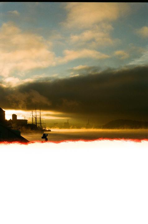 A ship is docked in a harbor with fog rolling over the water and buildings in the distance under a cloudy sky.