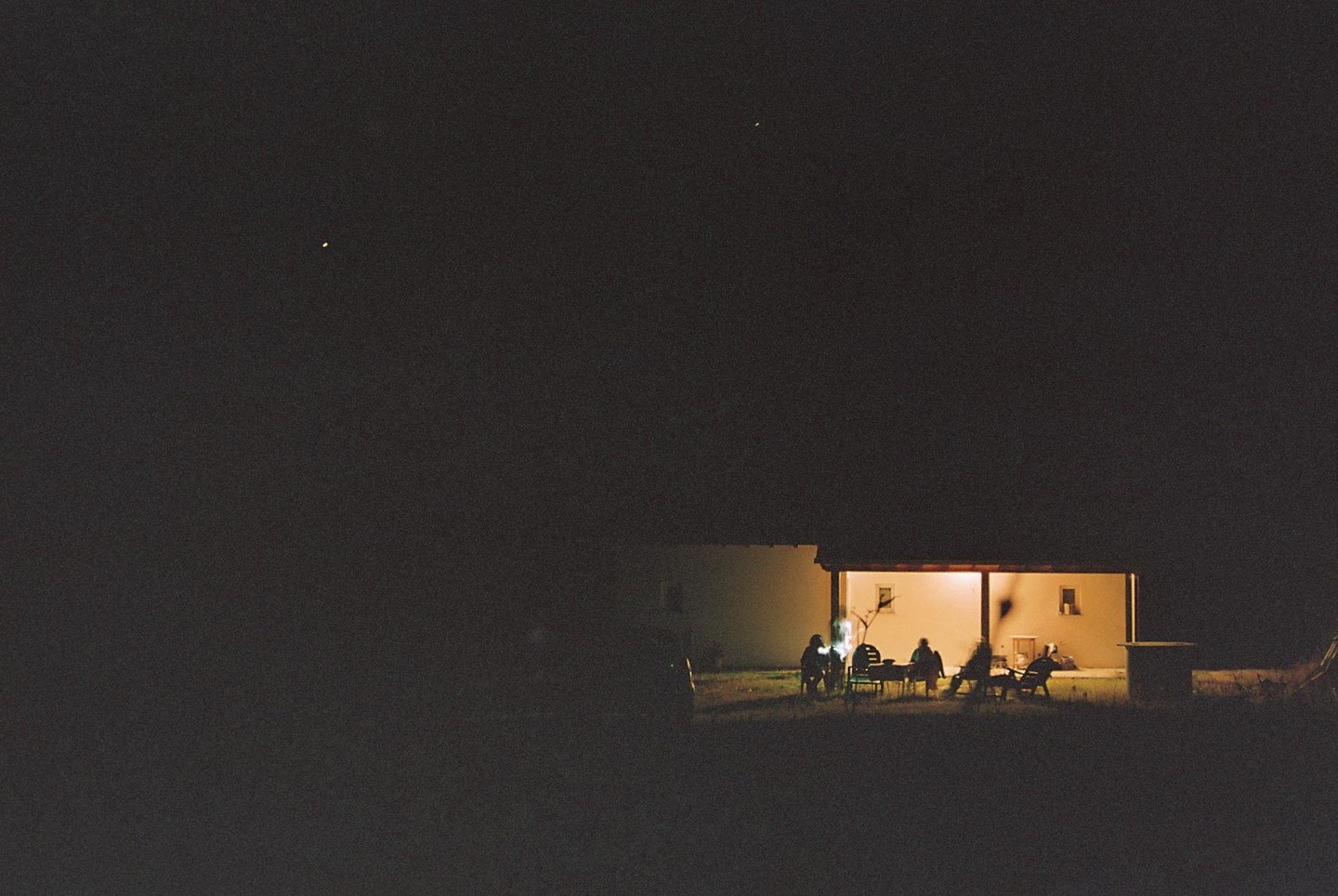 A group of people sit around a table on a porch at night, illuminated by a warm light.