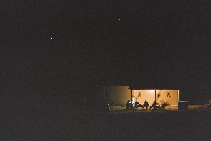 A group of people sit around a table on a porch at night, illuminated by a warm light.