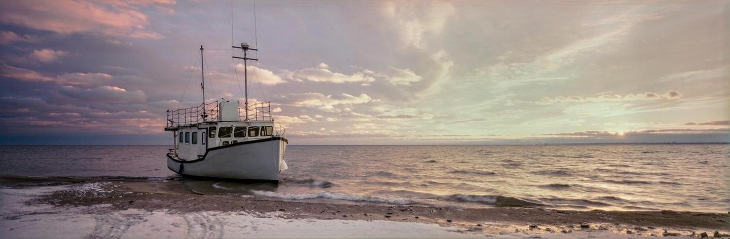 A white boat sits on a snowy beach next to the water under a cloudy sky at sunset.