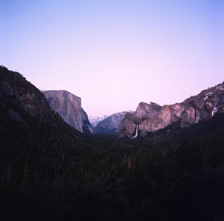 A wide valley with a waterfall and a forest is surrounded by mountains under a purple sky.