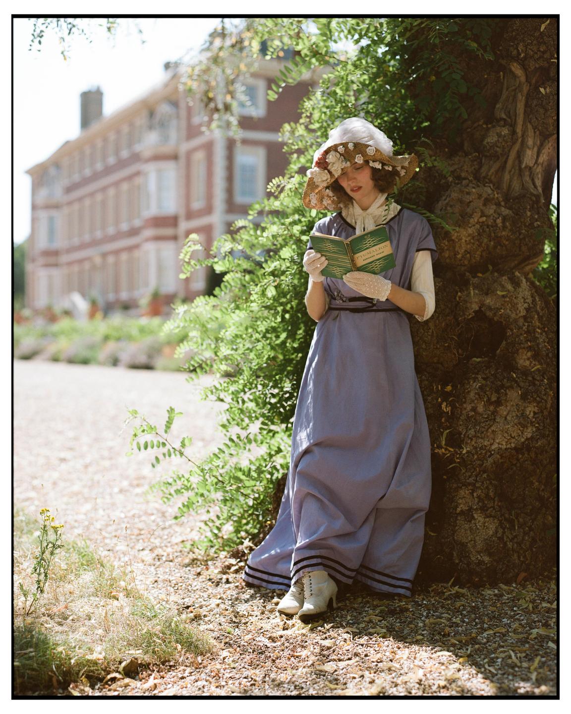 A woman in a period dress and a floral hat reads a book while leaning against a tree.