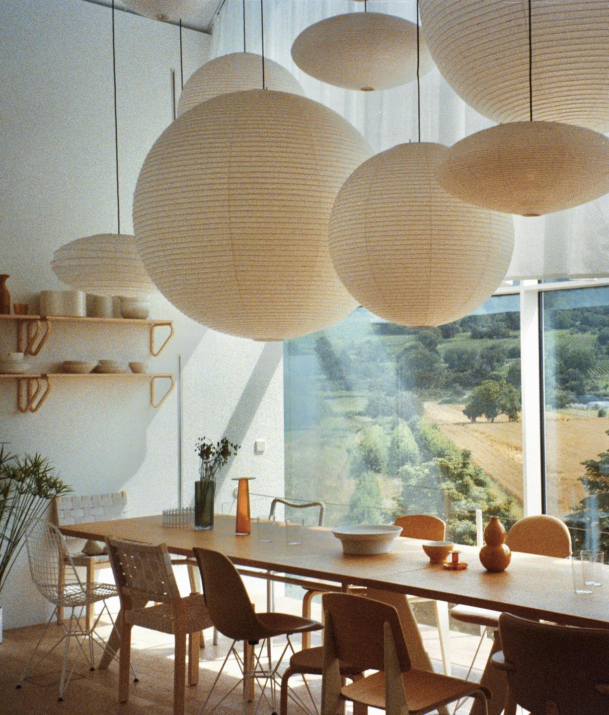A dining room with a long wooden table and several paper lanterns hanging from the ceiling overlooks a green landscape.
