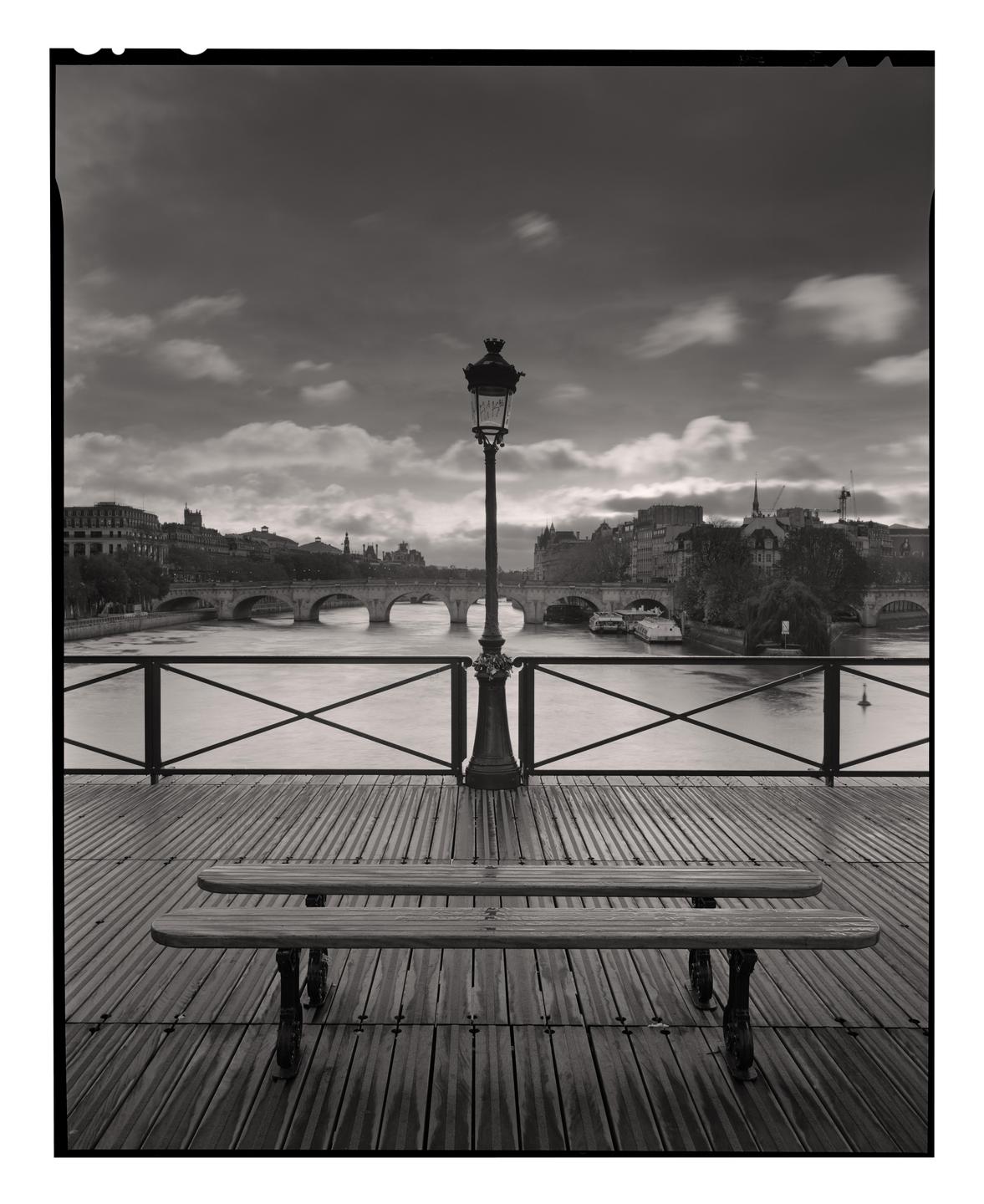 A lamppost and two benches are on a wooden deck overlooking a river with bridges and buildings in the distance under a cloudy sky.