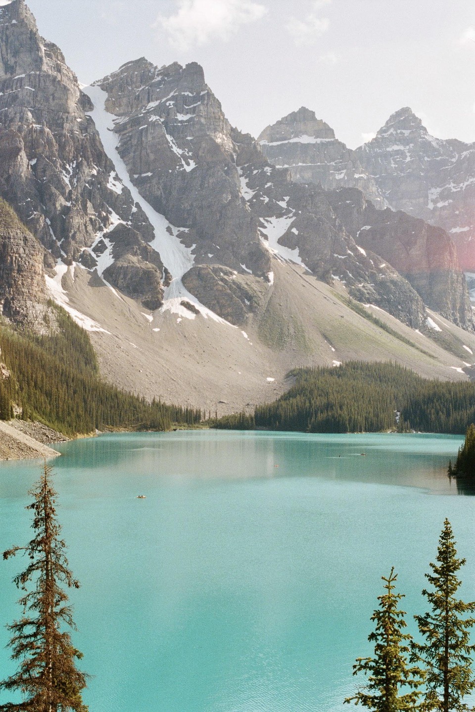 Moraine Lake is surrounded by snow-capped mountains and pine trees, with a few canoes visible on the turquoise water.