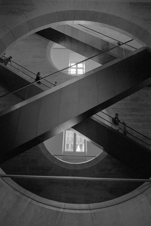 People ride escalators that crisscross within a circular architectural space, with windows visible in the background.