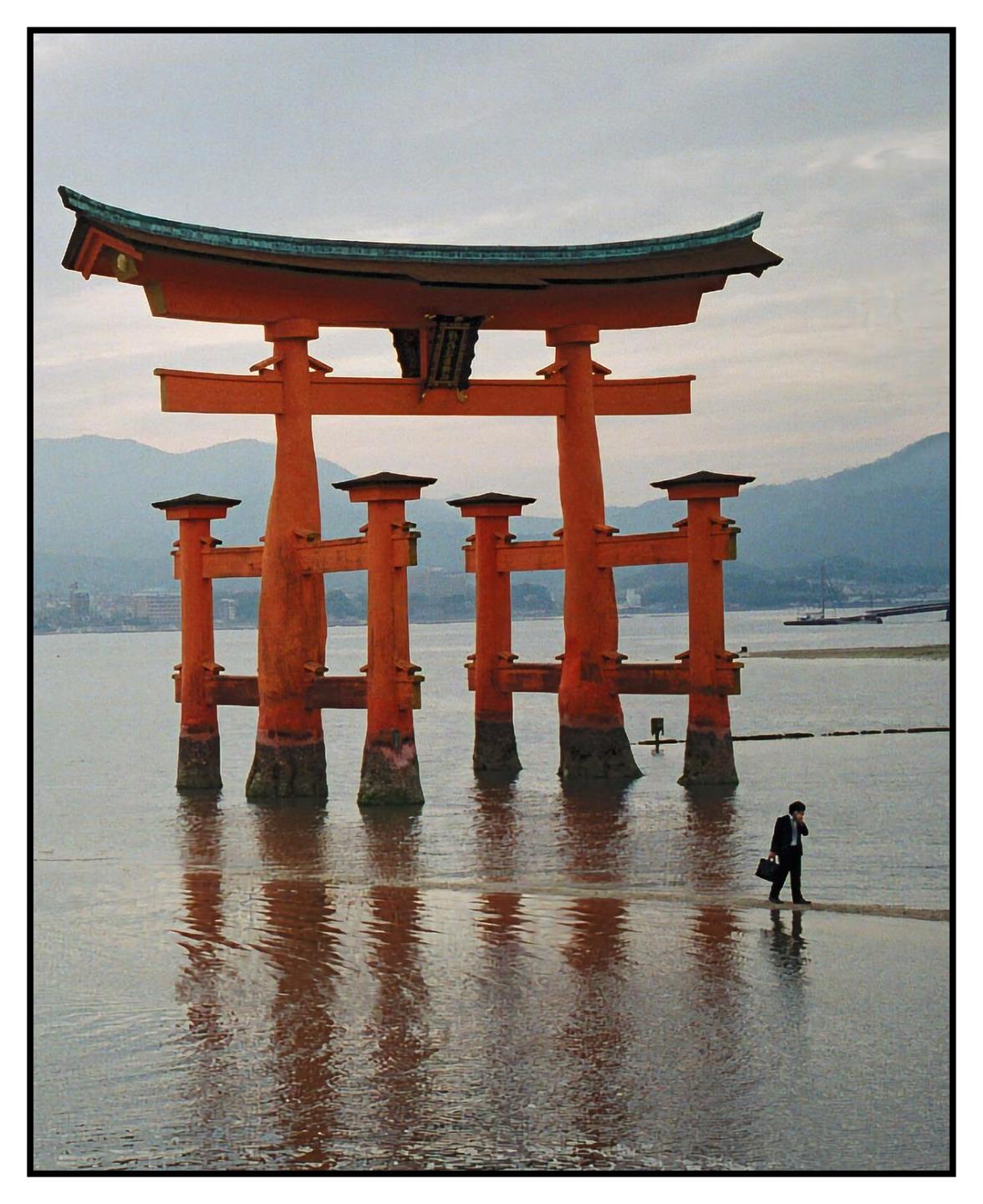 A man walks through shallow water near a large orange torii gate in the sea.