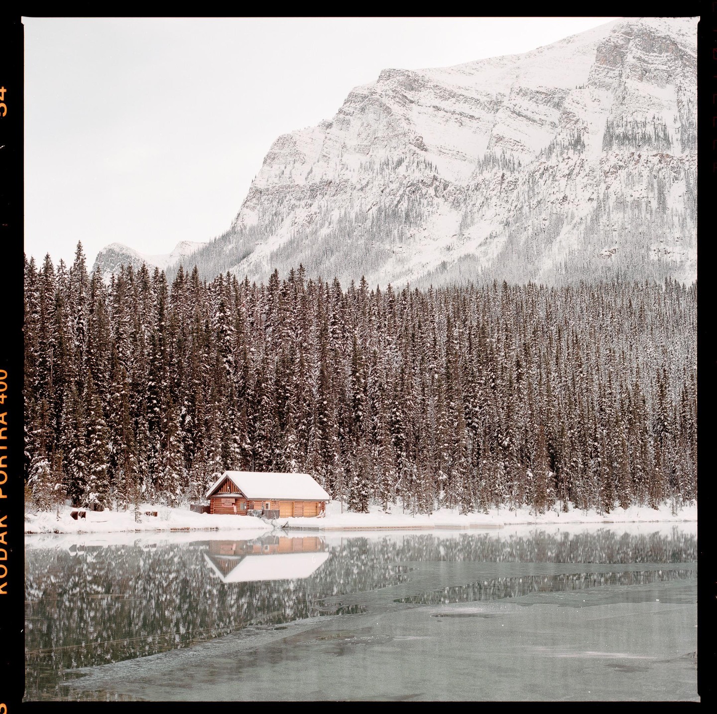 A wooden cabin sits by a partially frozen lake, surrounded by snow-covered pine trees and mountains.